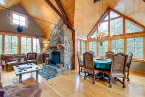 Un salón amplio con chimenea de piedra, vigas de madera en el techo y grandes ventanales con vistas al bosque.