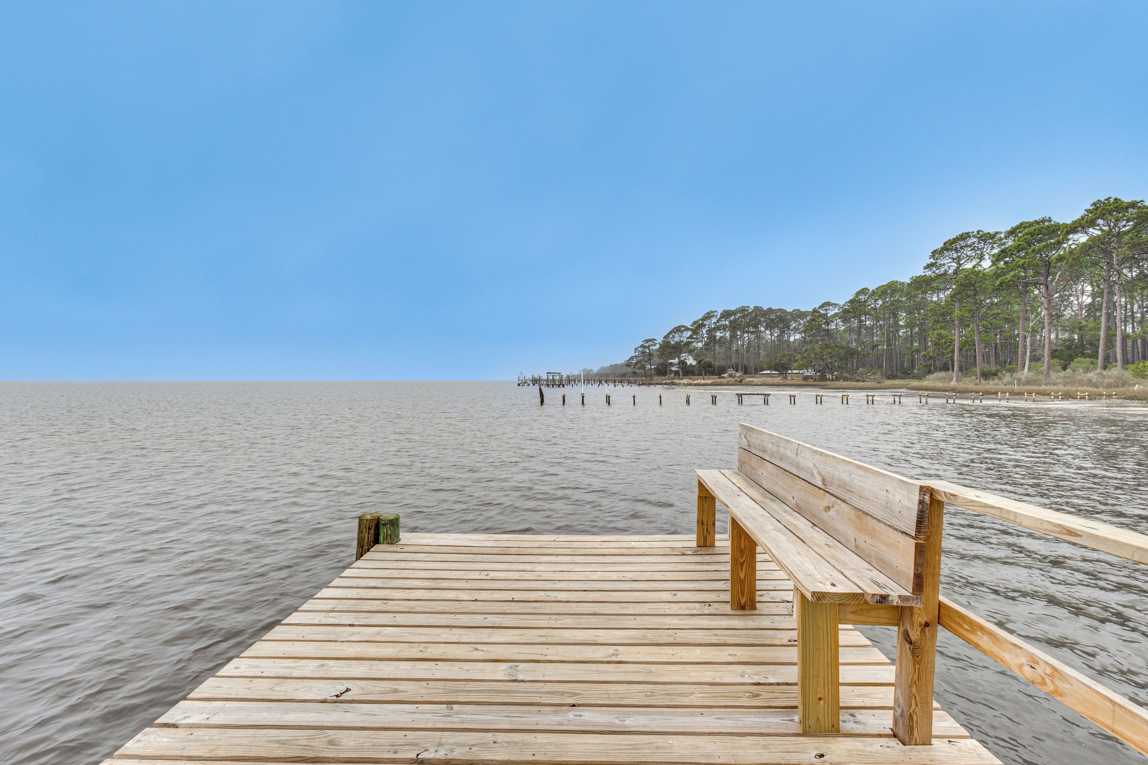 A wooden pier extending into calm water with a bench in the foreground and trees in the background.