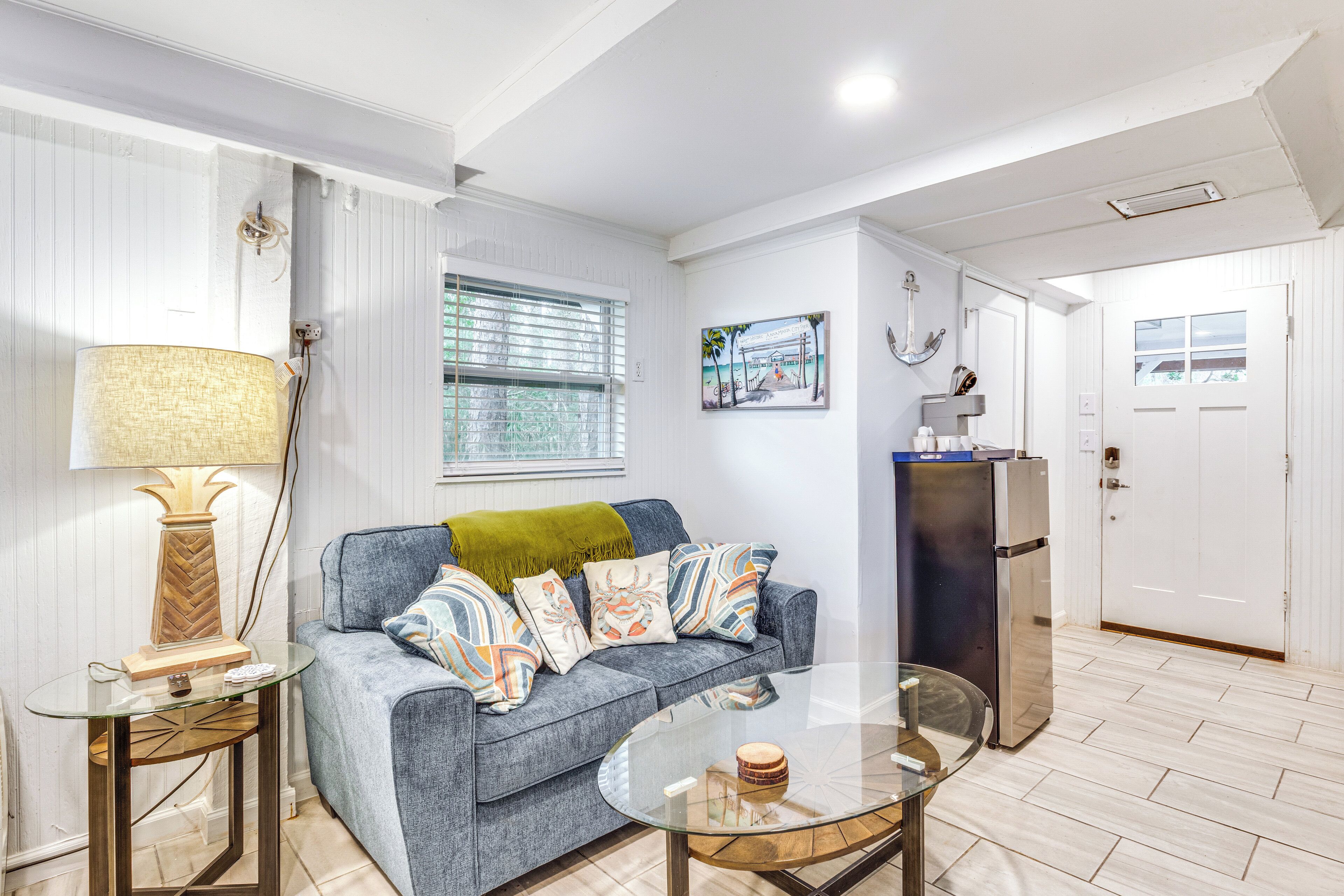A living room with a glass coffee table, a grey sofa with patterned cushions, a lamp, and a refrigerator.