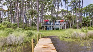 Un muelle de madera que lleva a una casa con techo rojo, rodeada de árboles altos y agua.