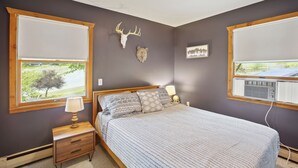 A bedroom with a bed, two windows, a nightstand, a lamp, a wall-mounted heater, and a wall decoration.