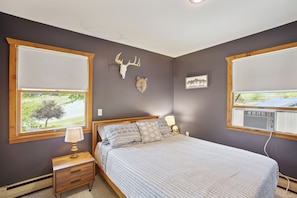 A bedroom with a bed, two windows, a nightstand, a lamp, a wall-mounted heater, and a wall decoration. - Crystal Lake Vacation Rental, 3 Mi to Betsie River (Beulah)