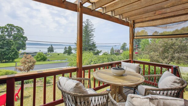 A covered outdoor seating area with wicker chairs and a table, overlooking a scenic landscape with trees and a body of water.