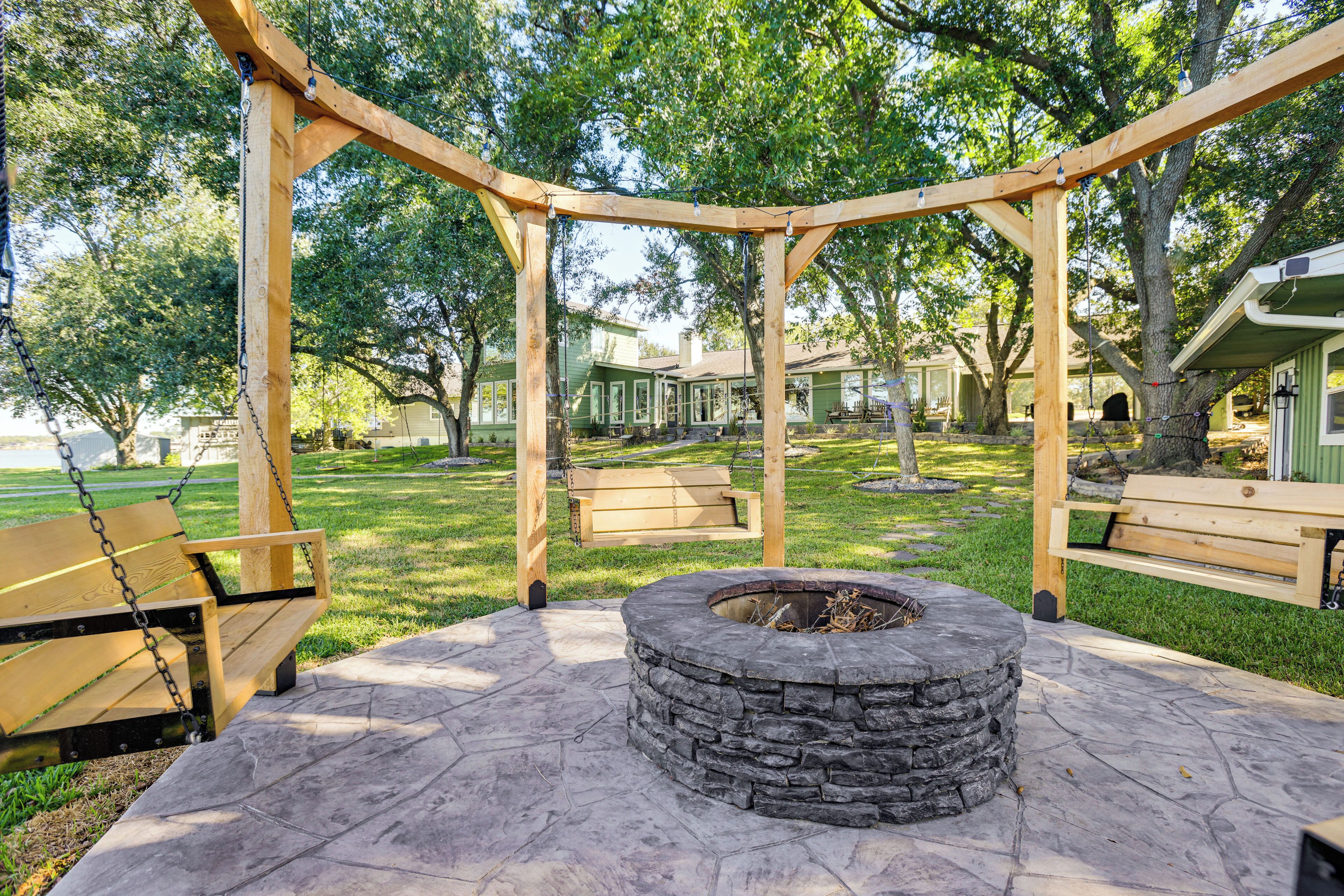 Un mirador con una fogata de piedra, bancos de madera y un conjunto de columpios en un parque con árboles y una casa al fondo.
