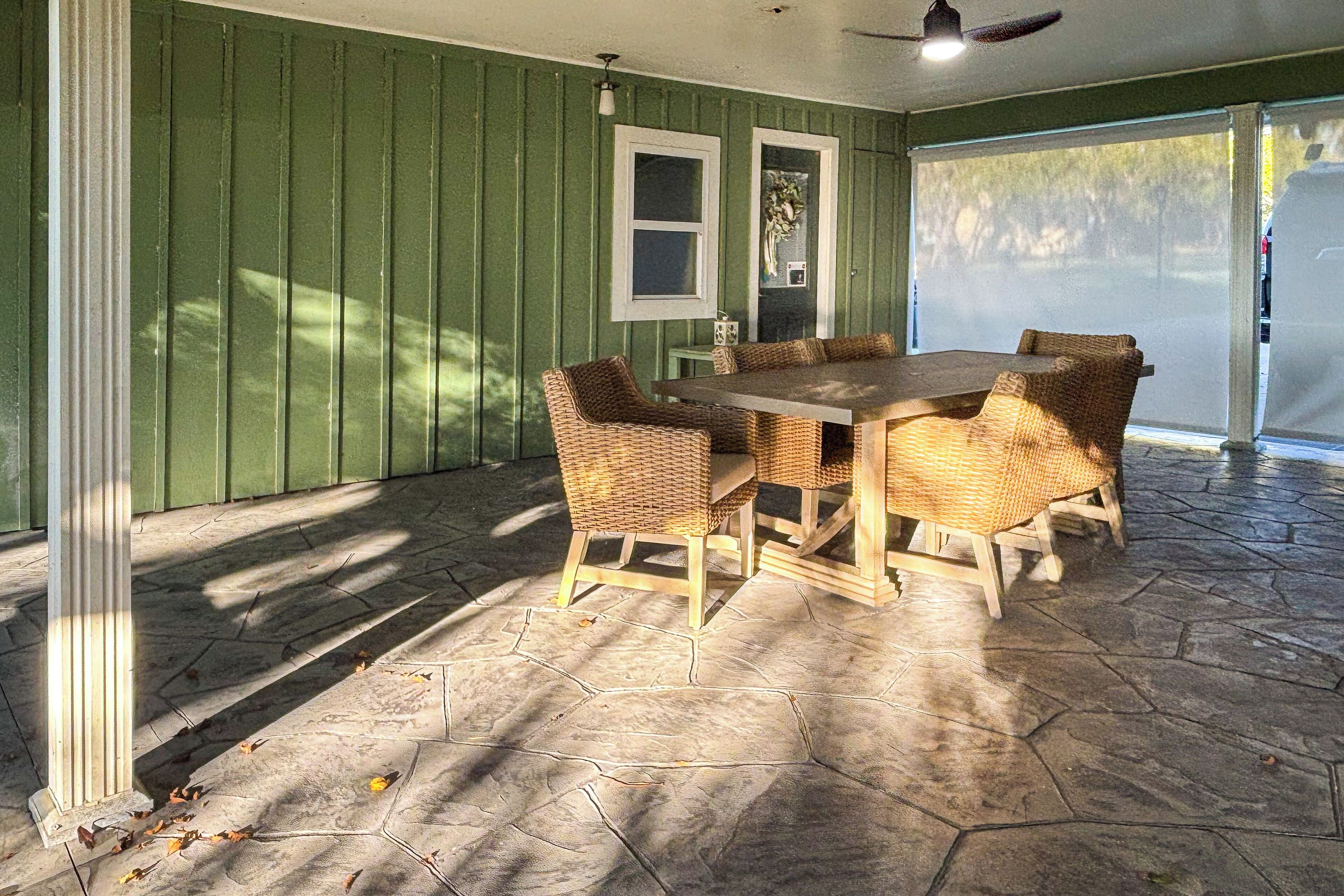 A covered patio with a hexagonal stone floor, a wooden table with wicker chairs, and a green paneled wall with two windows.