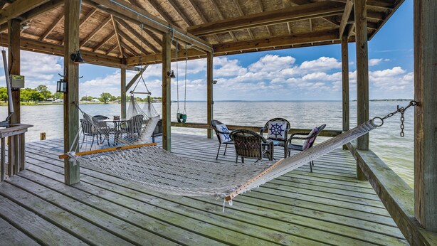 Eine Holzterrasse mit einer Hängematte, Korbstühlen und einem Tisch mit Blick auf ein Gewässer.