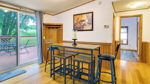 A dining area with a wooden table and four stools, a large window with a view of trees, and a painting on the wall. - Charming Watkins Glen Home w/ Deck & Yard! (Watkins Glen)