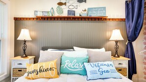 A bedroom with a bed, two bedside tables, lamps, decorative pillows, a shelf with items, and a curtain. - Cozy Cottage: Minutes to Downtown Southport! (Southport)