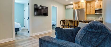 A modern living room with a gray sofa, wooden dining table, and kitchen area with wooden cabinets and stainless steel appliances.