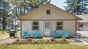 Een klein huis met een veranda, een barbecue en drie blauwe Adirondack-stoelen.