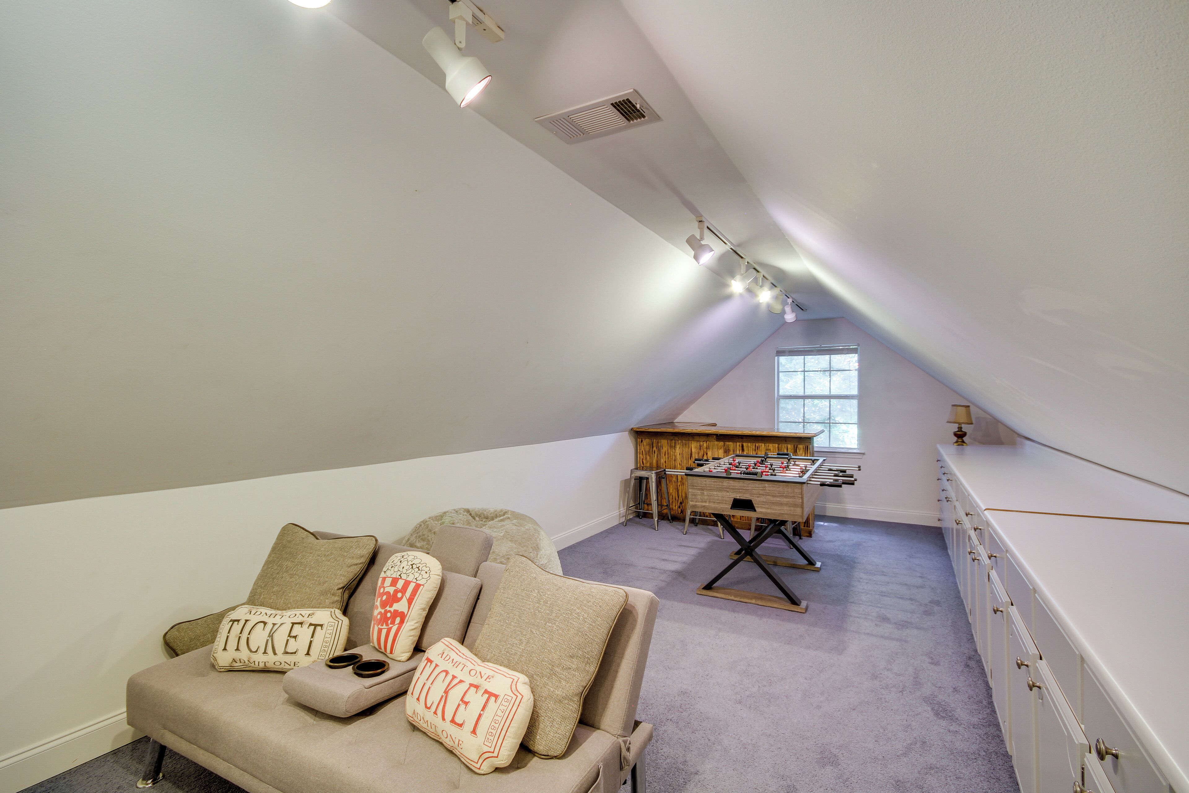 Une chambre à coucher confortable aménagée en mezzanine, avec un plafond en pente, un canapé agrémenté de coussins sur le thème des « billets », une table de billard et une grande fenêtre.