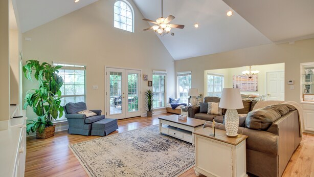 A spacious living room with a high ceiling, large windows, and a ceiling fan. - Charming Diamondhead Home w/ Private Yard! (Diamondhead)