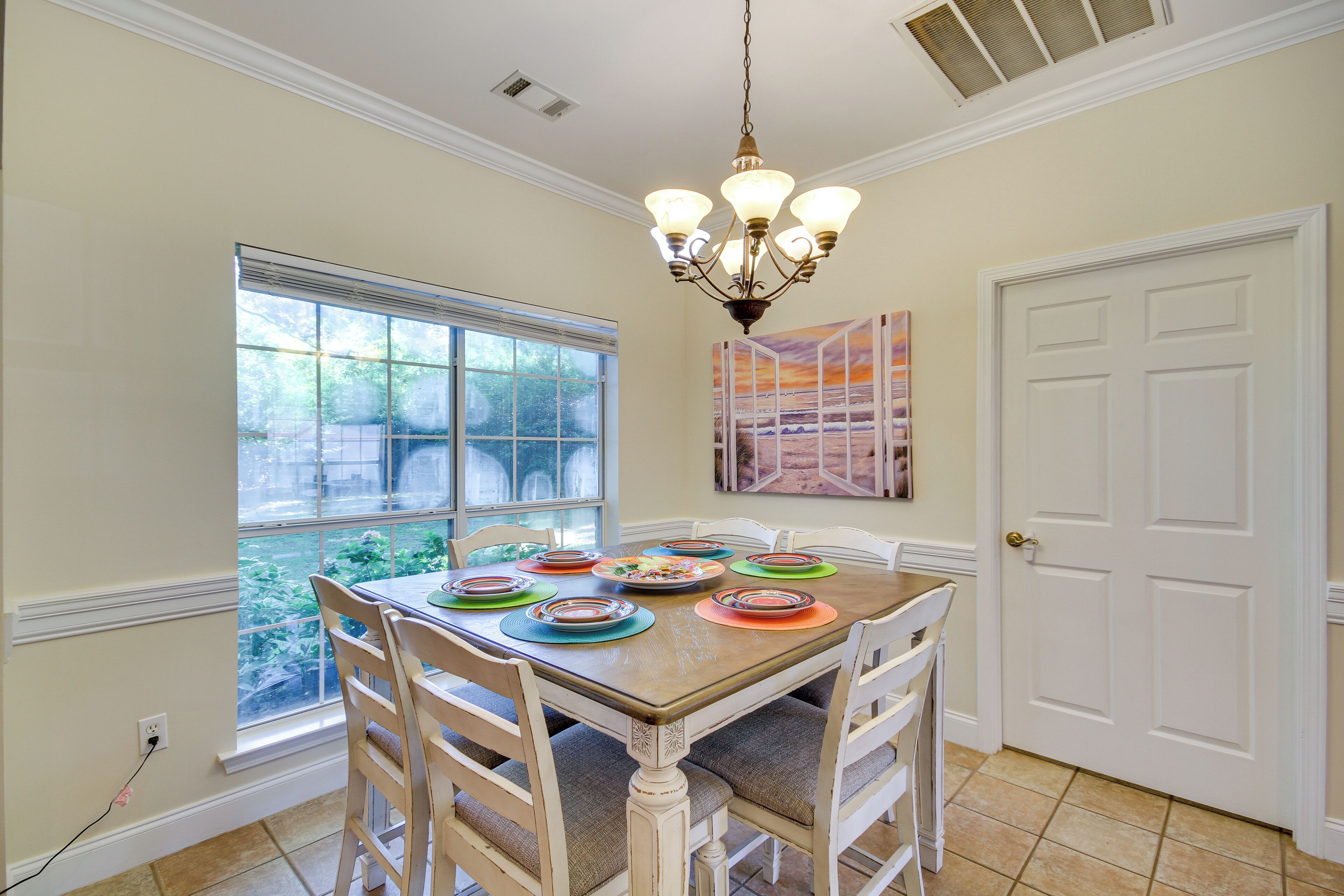 A dining room with a wooden table set for six, surrounded by white chairs. A chandelier hangs from the ceiling, and a window provides a view of greenery outside.