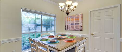 A dining room with a wooden table set for six, surrounded by white chairs. A chandelier hangs from the ceiling, and a window provides a view of greenery outside.