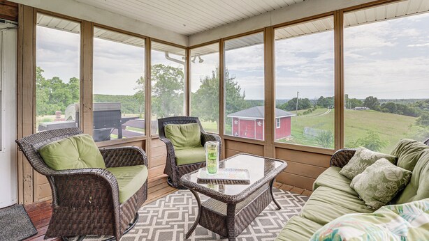 Een overdekte veranda met rieten meubels, een glazen tafel en uitzicht op een rode schuur en groen landschap.