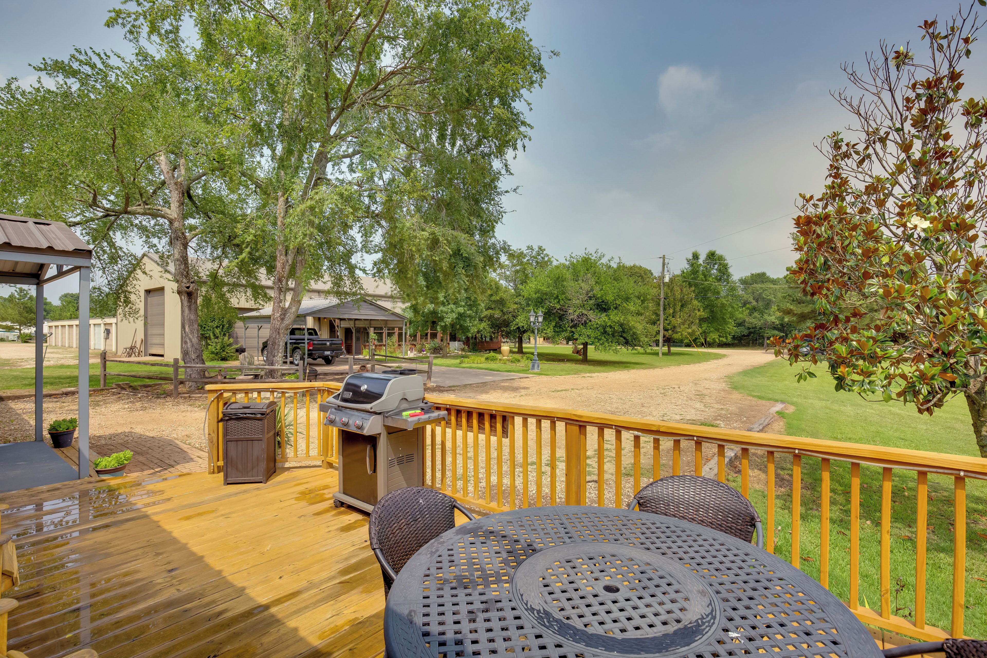 Une terrasse en bois, meublée pour l’extérieur, avec un barbecue et une vue sur une zone herbeuse et des arbres.