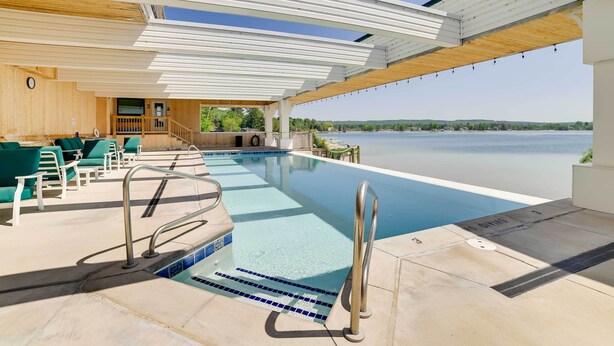 Zona de piscina con vista a un lago, un muelle de madera y un área de estar cubierta.