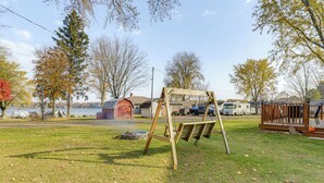 Une zone herbeuse avec une balançoire en bois, une structure de jeux couverte et, en arrière-plan, une vue sur un lac et des camping-cars.