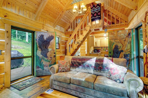 Ein gemütliches Blockhaus-Interieur mit Holzbalkendecke, einer Couch mit Decke und einer Treppe, die zu einer oberen Ebene führt.