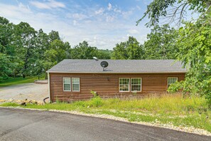 A single-story log cabin with a satellite dish, surrounded by trees and a gravel driveway.