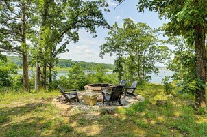 A fire pit with chairs and a cooler in a wooded area with a lake view.
