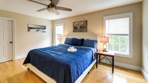 Une chambre à coucher comprenant un lit, un ventilateur de plafond, deux fenêtres équipées de stores, une table de chevet avec une lampe et des œuvres d’art accrochées aux murs.