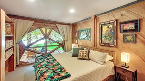 A bedroom with a large window, wooden walls, a bed with a quilt, pillows, a bedside table with a lamp, and framed artwork on the wall. - Unique Nordman Retreat 1 Mi to Priest Lake! (Nordman)