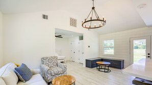 A modern living room with a sofa, patterned armchair, and round coffee table. A kitchen area with dark cabinetry and a dining table is visible. - Bright Wallis Cottage w/ Pergola & Grill! (Wallis)