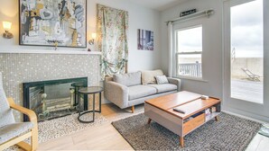 Ein modernes Wohnzimmer mit Kamin, einem grauen Sofa, einem Couchtisch und einem großen Fenster mit Blick auf den Strand.