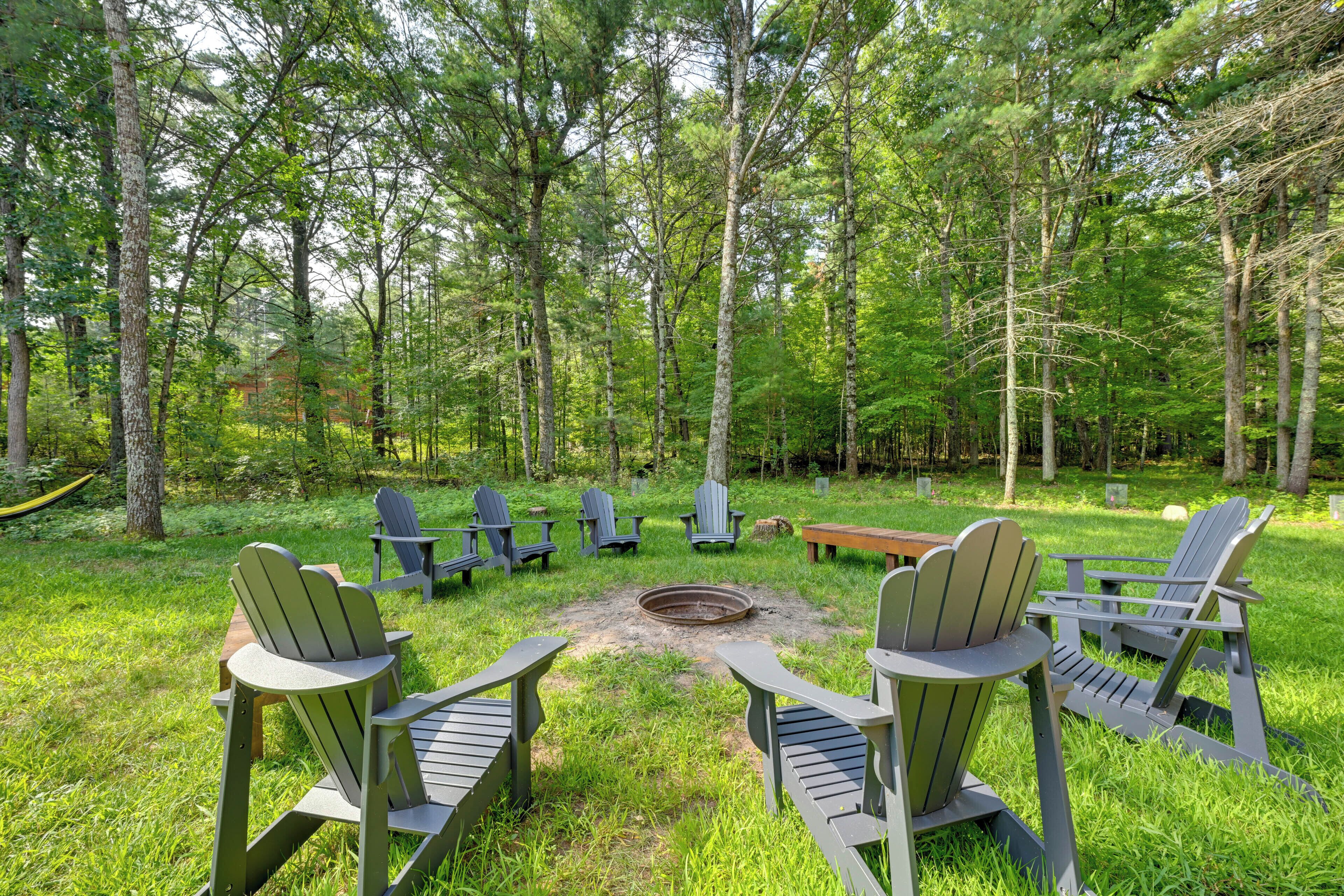 A forested area with Adirondack chairs arranged around a central fire pit.