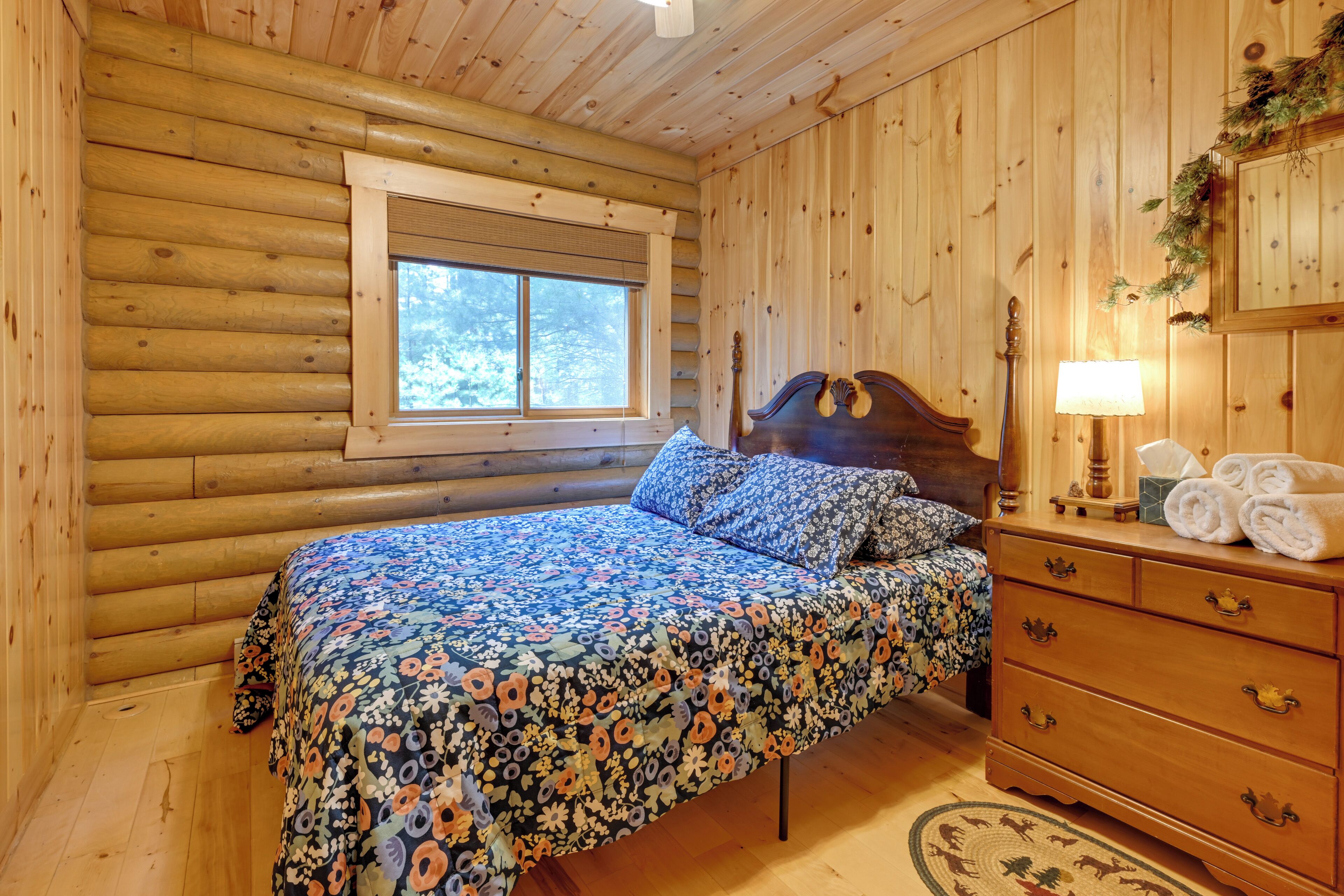A log cabin bedroom with a wooden bed, a dresser with towels, and a window with blinds.