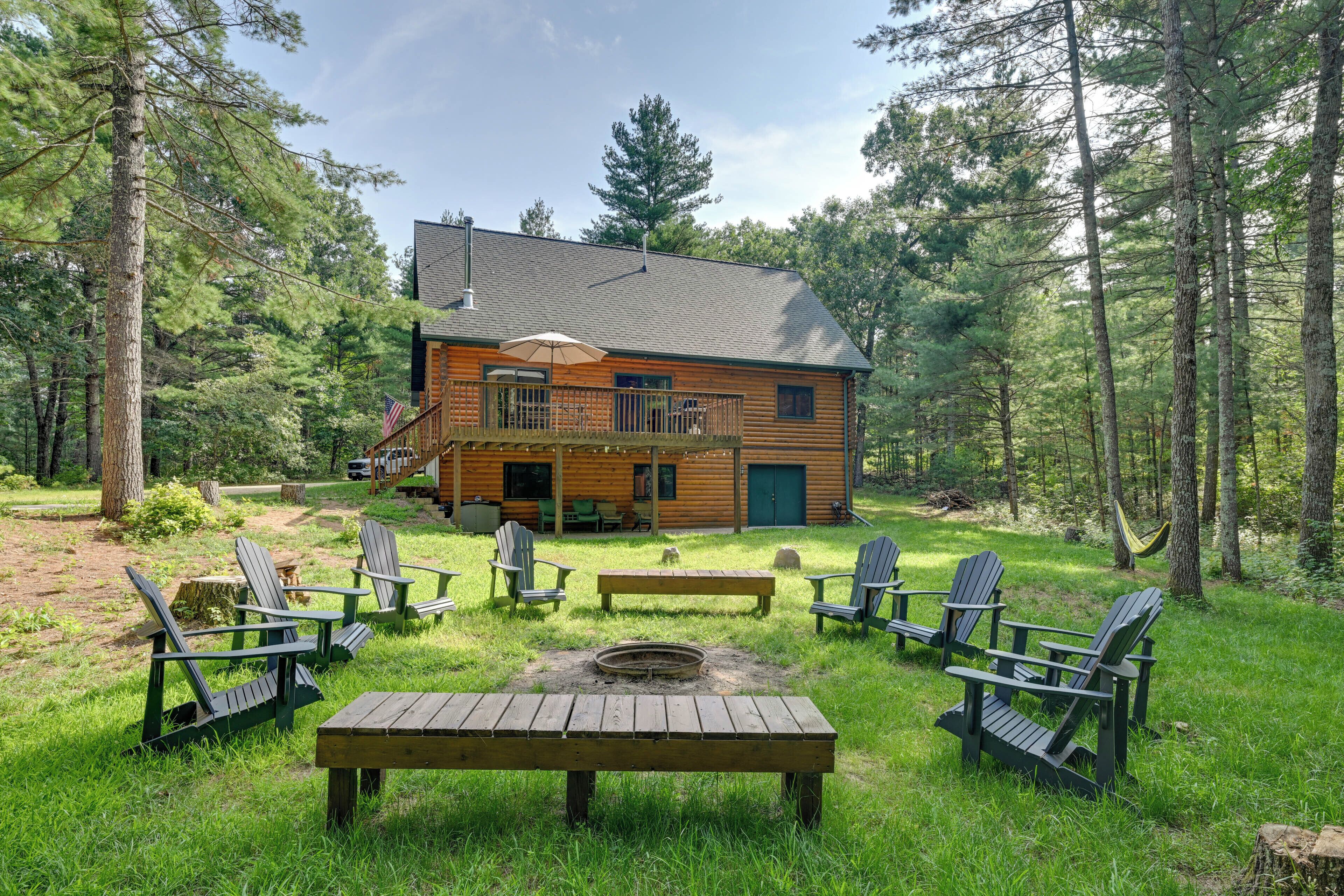 A cabin with a deck, surrounded by a forest. There are Adirondack chairs, a fire pit, and a picnic table.