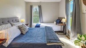 Un dormitorio con dos camas, un ventilador de techo, una ventana con cortinas, una mesita de noche con lámpara y una planta en maceta.