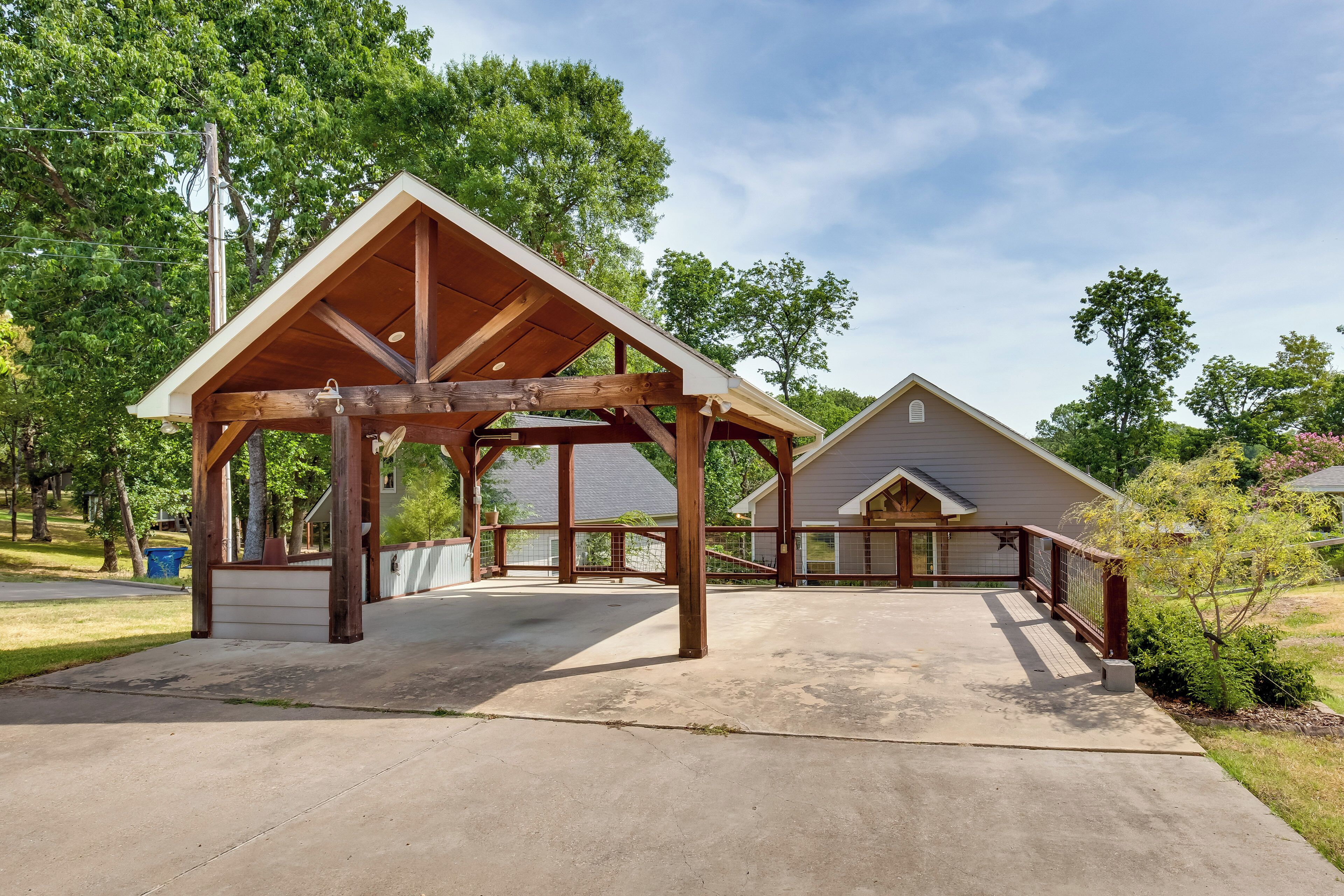 A covered outdoor area with a wooden structure, a concrete floor, and a view of a house in the background.