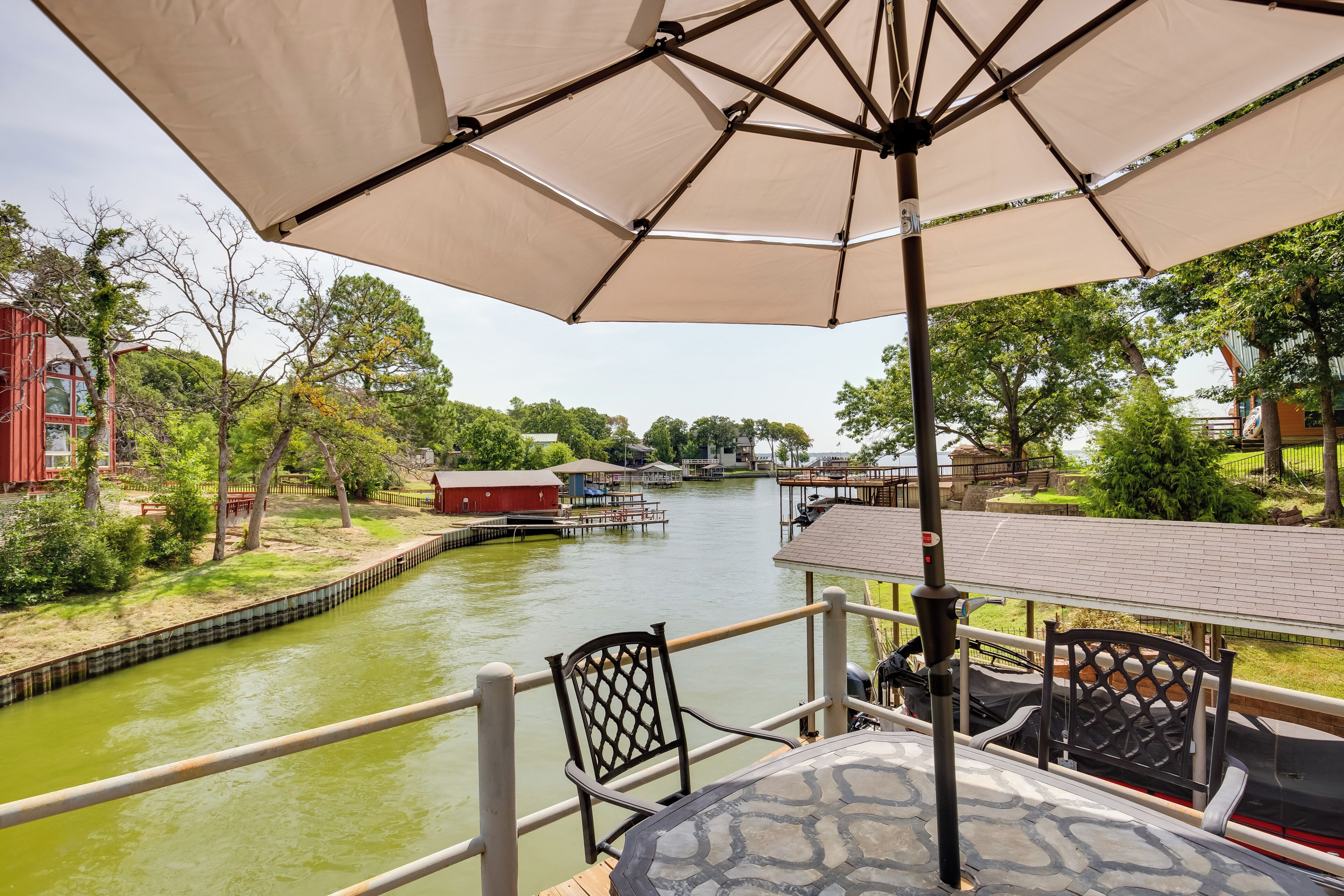 A riverside patio with a table and chairs under a large umbrella, overlooking a canal with boats and buildings in the background.