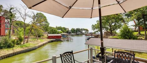 A riverside patio with a table and chairs under a large umbrella, overlooking a canal with boats and buildings in the background.