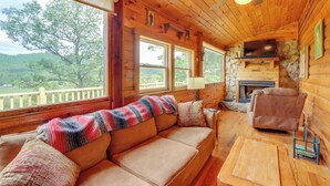 A cozy log cabin interior with a wooden ceiling, a stone fireplace, a sofa with a blanket, and a television. - Secluded Mountain Cabin w/ Decks + Gazebo! (Warne)