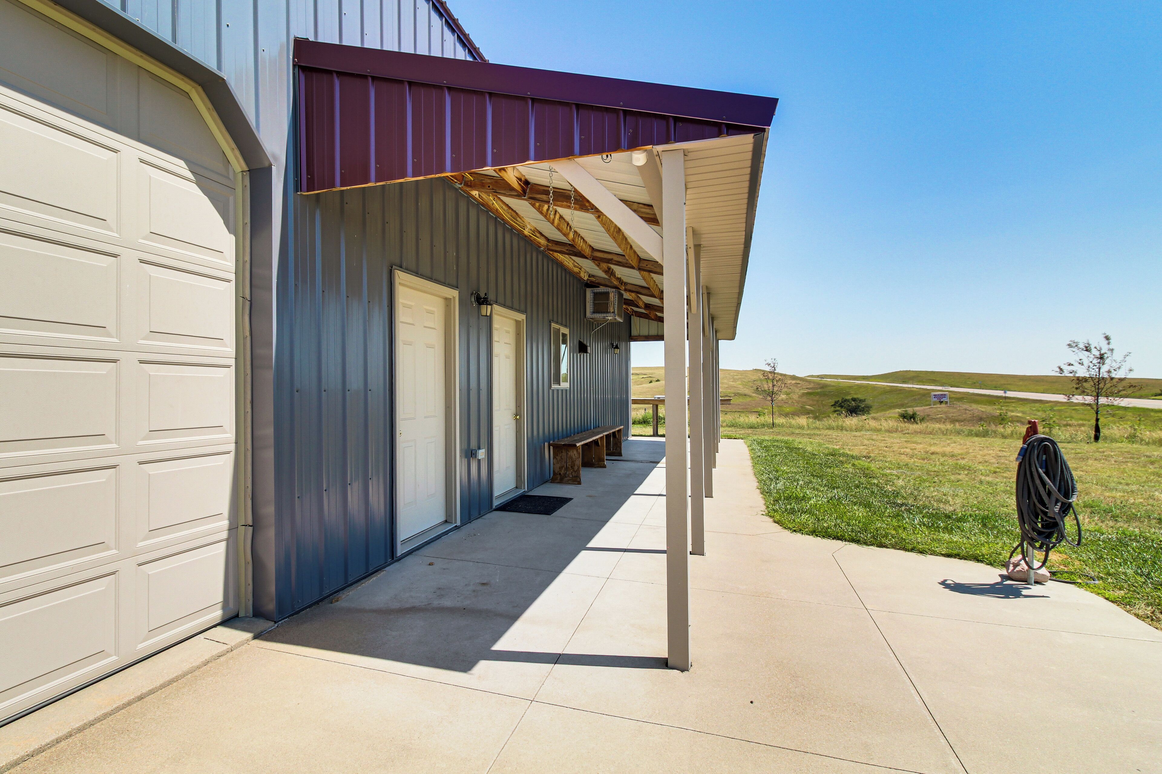 A covered porch with a metal roof, a concrete walkway, and a grassy field in the background.