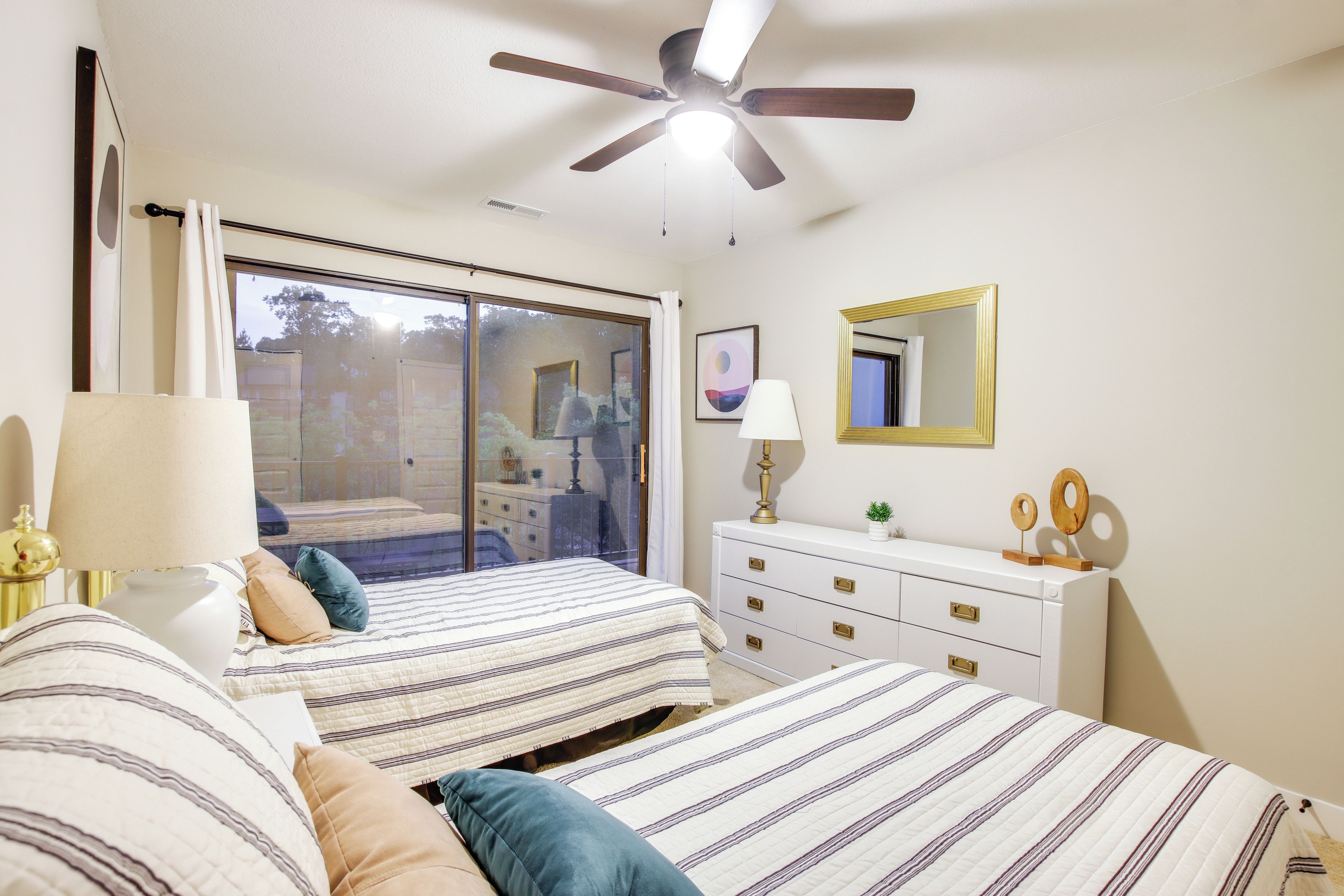 A bedroom with two beds, a ceiling fan, a dresser, a mirror, and a sliding glass door leading to a balcony.