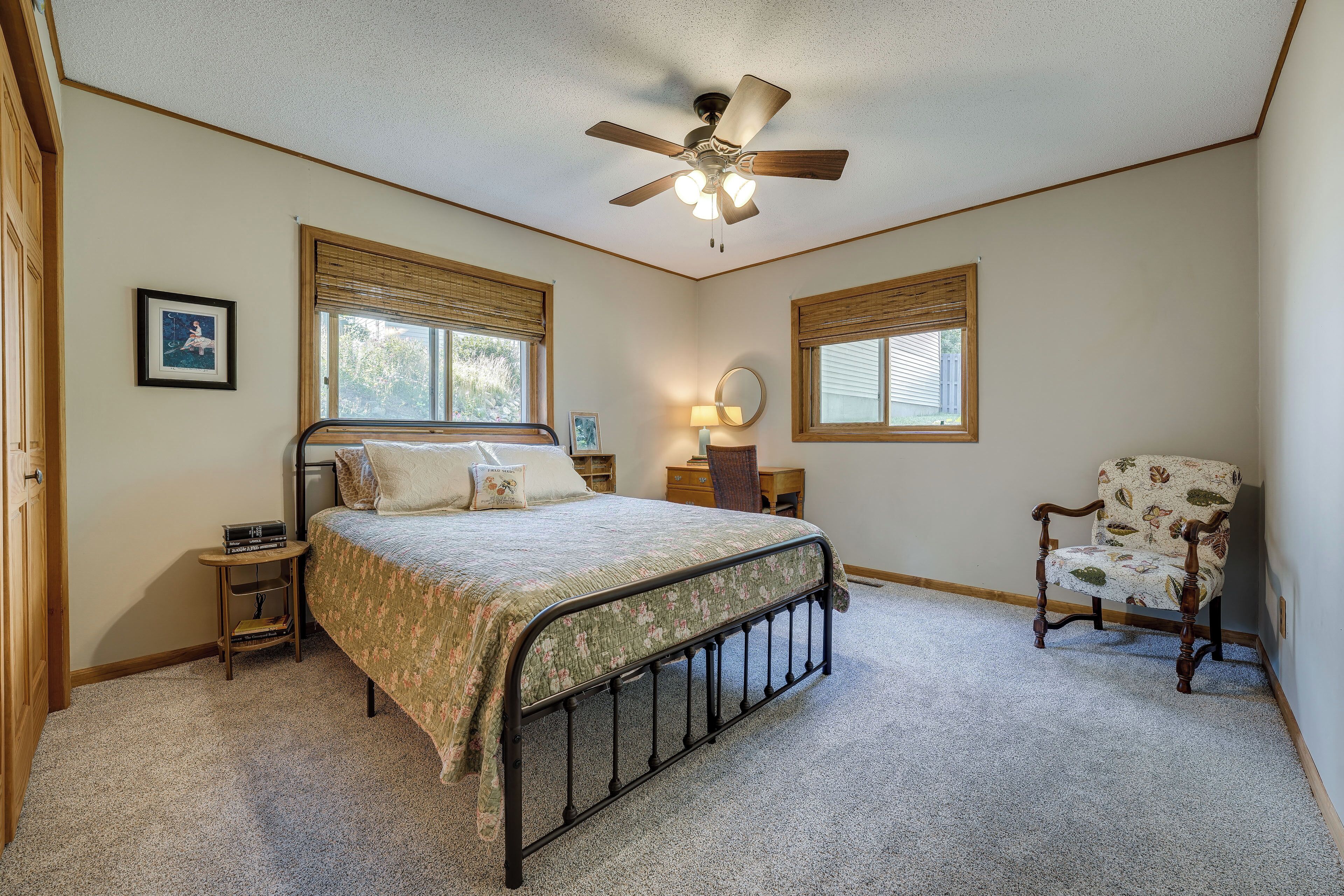 A bedroom with a bed, a chair, a desk, a ceiling fan, and two windows with blinds.