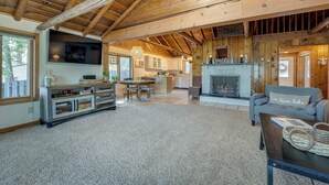 A spacious living room with a fireplace, wooden ceiling beams, a dining area, and a kitchen in the background. - Delton Oasis Retreat w/ Lake Views & Deck! (Delton)