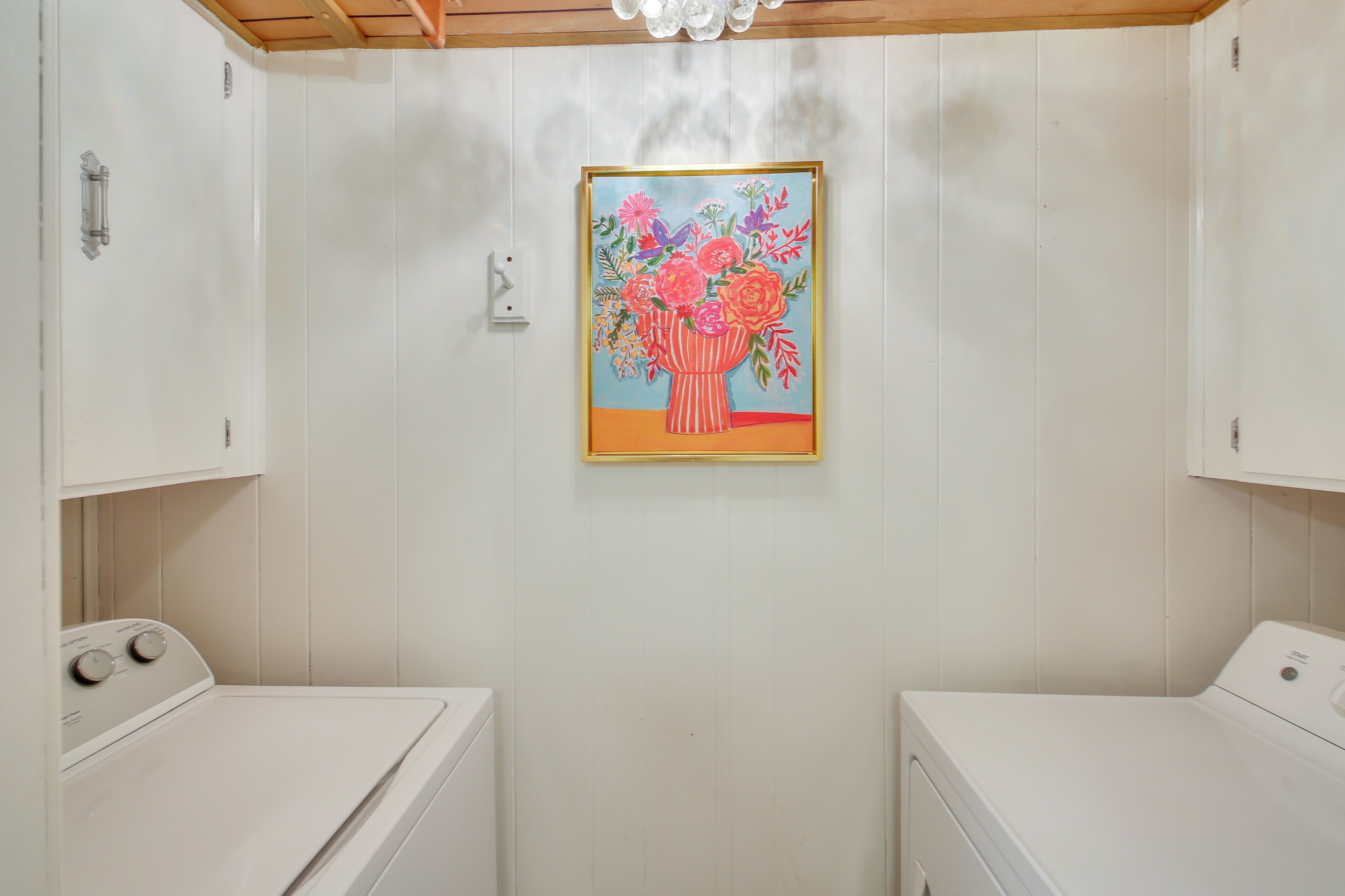 A laundry room with two washing machines, a white wall, and a framed painting of flowers.