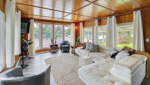 Un salon spacieux avec un plafond en bois, de grandes fenêtres, un canapé blanc, un fauteuil gris et un téléviseur.