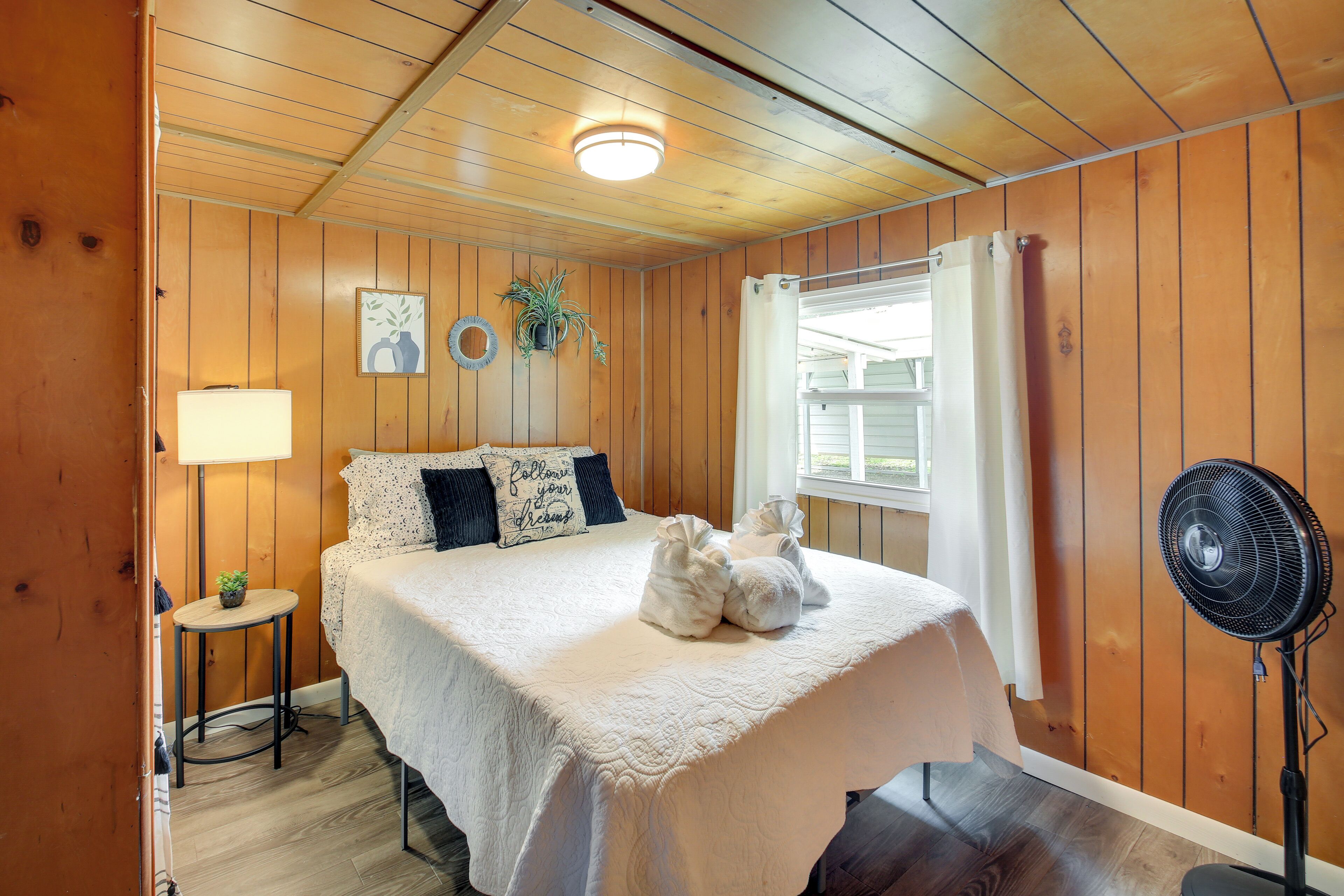Un dormitorio con paneles de madera, una cama, una mesita de noche, una lámpara y un ventilador de pie.