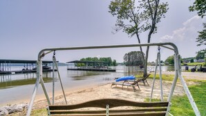 A wooden swing chair with metal supports, a sandy beach, a blue lounge chair, and a dock extending into a calm body of water. - Chic Spring City Townhome on Watts Bar Lake! (Spring City)