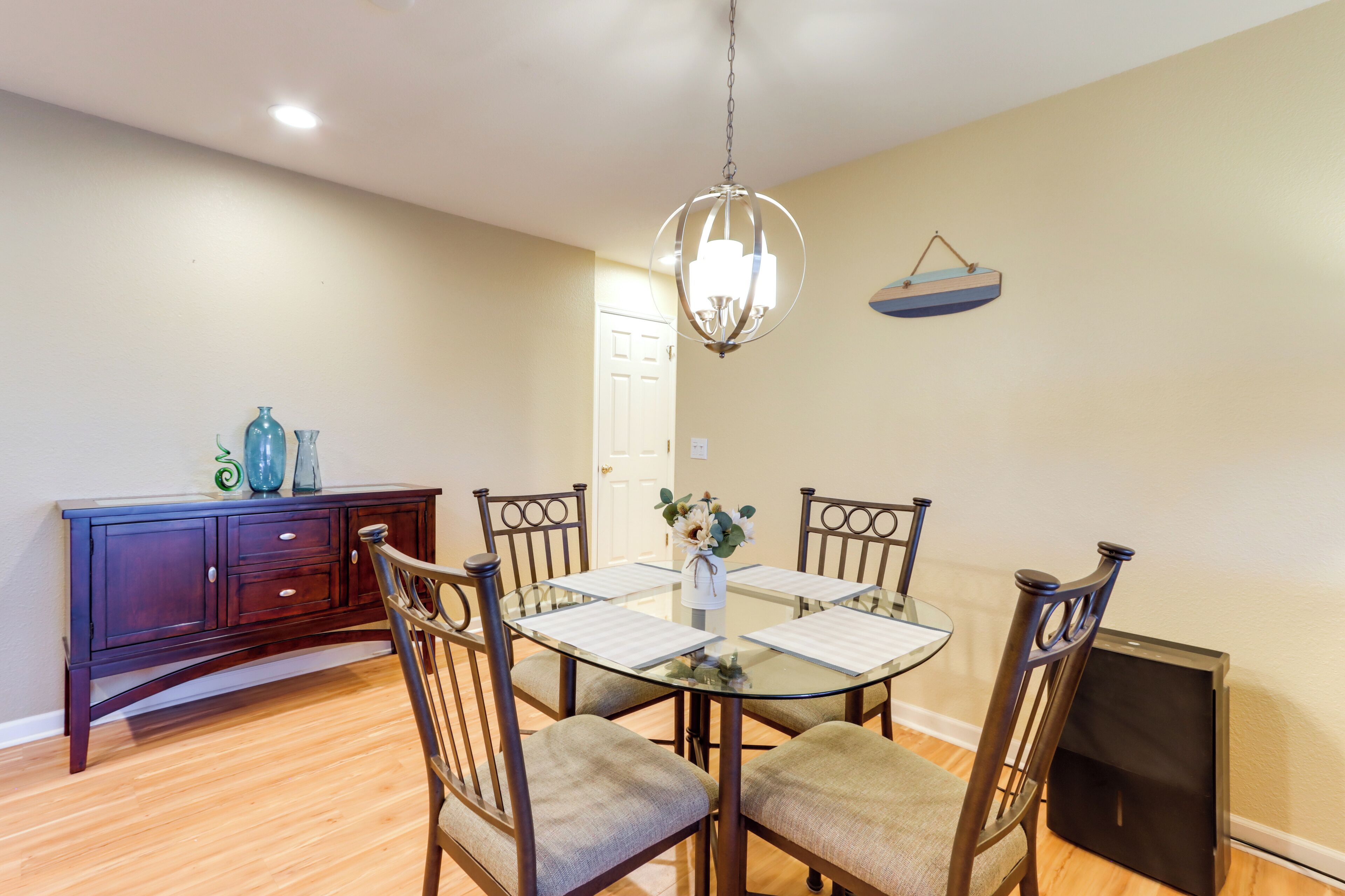 A dining area with a glass table, four chairs, a chandelier, a dark wooden cabinet, and a small refrigerator.