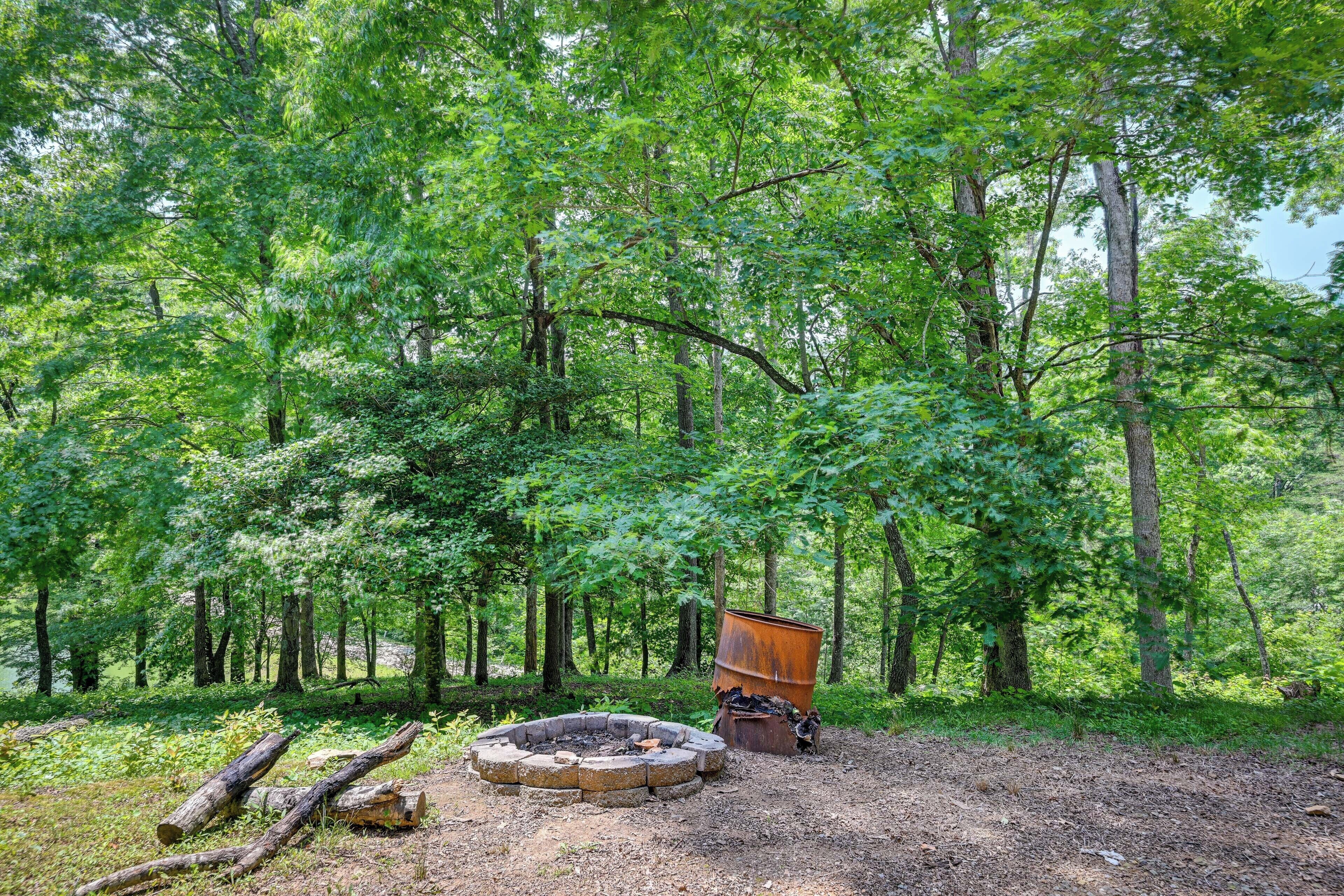 A forested campsite with a circular fire pit and a rusty barrel.
