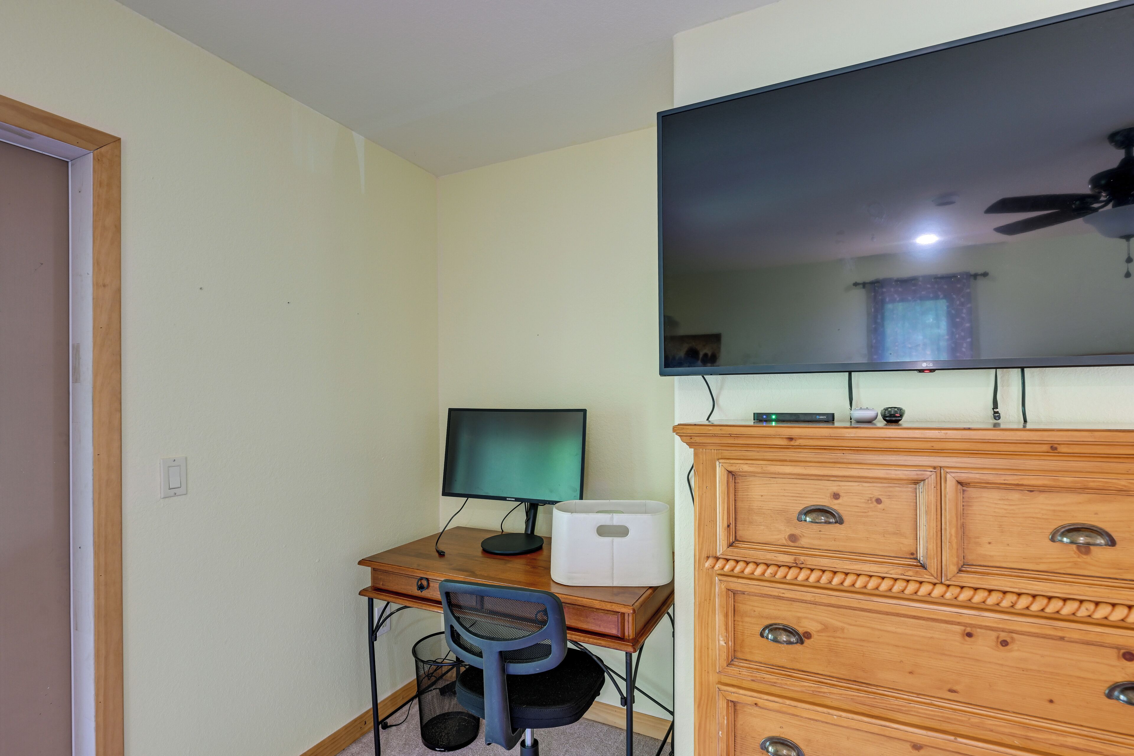 Une pièce comprenant un bureau en bois, un écran d’ordinateur, une chaise, une commode et un ventilateur de plafond.