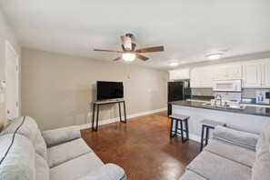 A modern living room with a ceiling fan, a flat-screen TV on a stand, and a kitchen area with white cabinets and a microwave. - Lovely Lake Charles Duplex in Central Location! (Lake Charles)