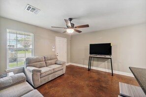 A living room with a beige couch, a television on a stand, a ceiling fan, and a window with blinds. - Lovely Lake Charles Duplex in Central Location! (Lake Charles)